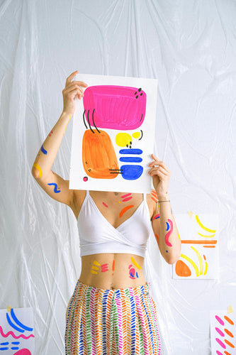 Woman stained with paint, holding up a colorful painting so that it covers up her face